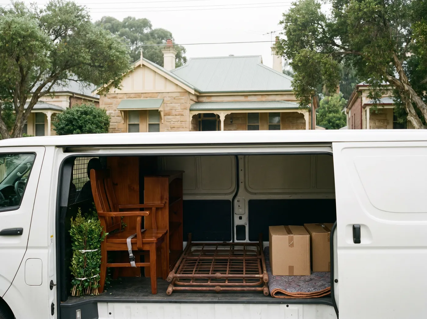 The open rear of a plain white van packed in tidy separated sections: strapped timber chairs and a small bookshelf, a tied bundle of hedge trimmings, a folded rusted iron bed frame and taped cartons, a leafy street of Federation houses behind